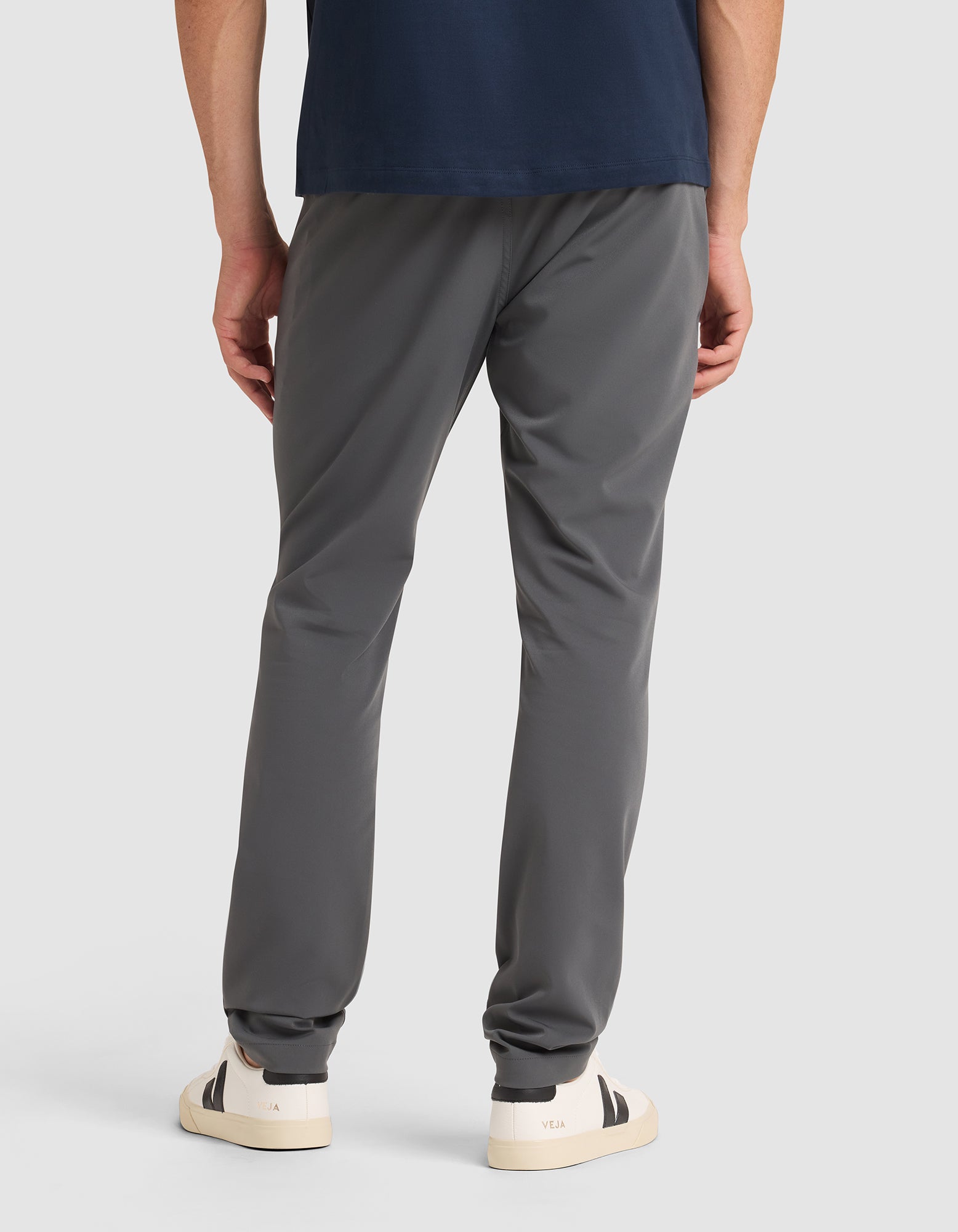 A person stands facing away, wearing Cozy Earth's Men's Everywhere Pant in dark gray, a navy blue shirt, and white sneakers with black heel accents against a plain light background.  