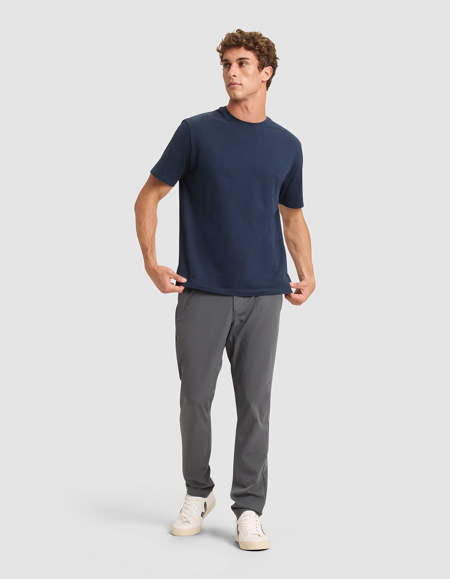 A man stands against a light gray background, wearing Cozy Earth's Men's Everywhere Pant in gray, paired with a plain navy blue T-shirt and white sneakers. He looks slightly to the side with a relaxed posture, one hand in his pocket.  