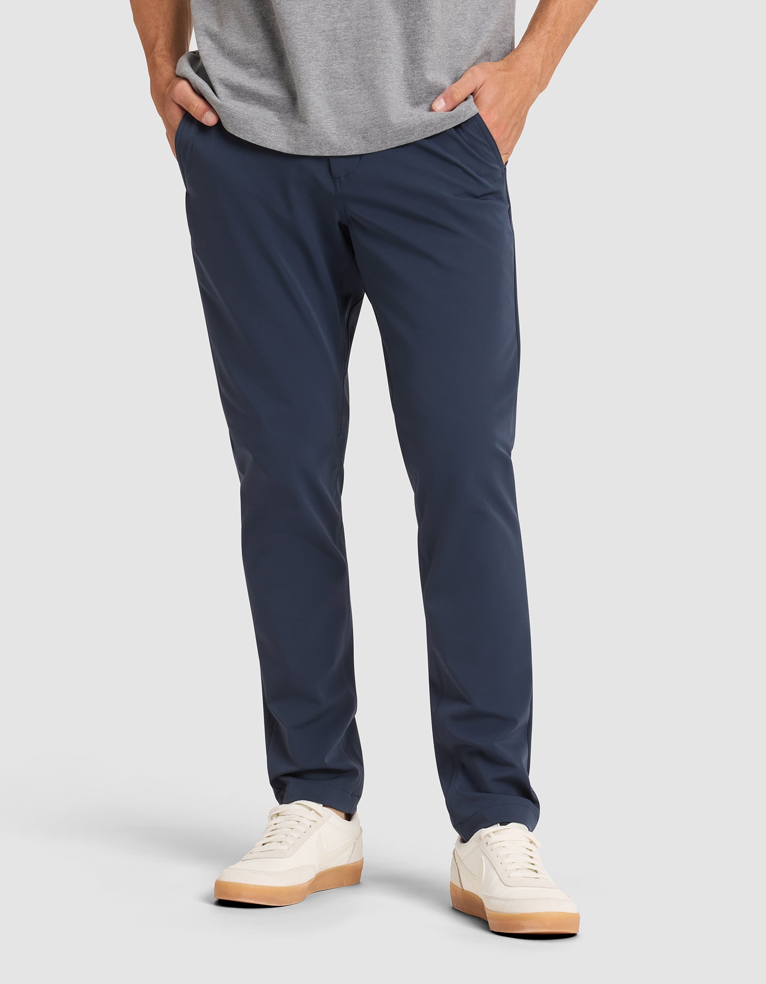 A person stands with hands in pockets against a plain background, wearing Cozy Earth's Men's Everywhere Pant in navy blue, a gray shirt, and white sneakers with light brown soles.  