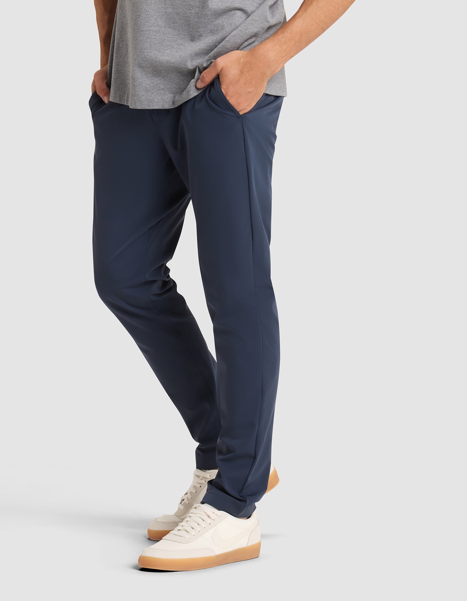 A person wearing Cozy Earth's Men's Everywhere Pant in navy, a gray tee, and white sneakers with gum soles stands hands-in-pockets against a light gray background. Only the lower body is shown. 