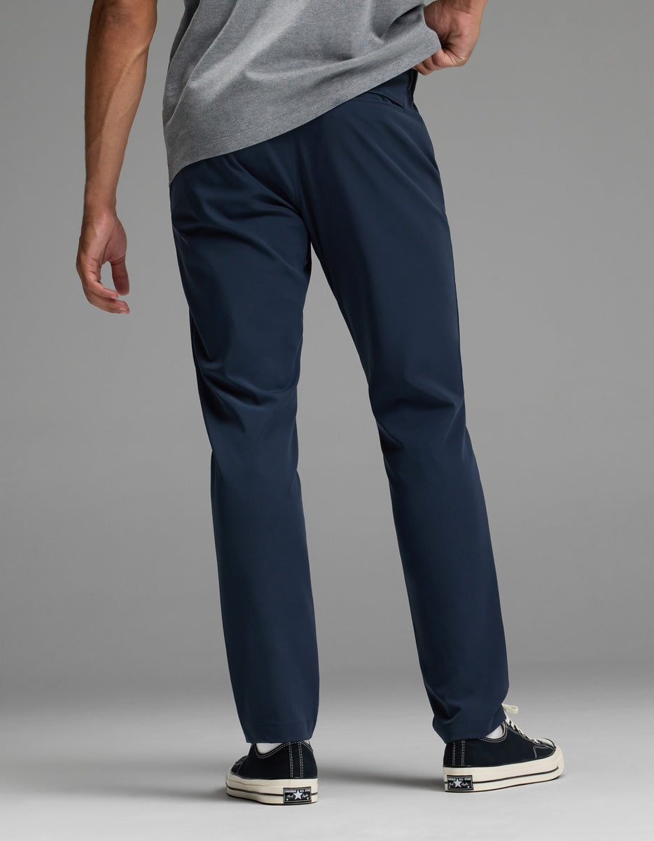 A man stands facing away against a plain background, wearing the Cozy Earth Men’s Everywhere Pant in navy blue, paired with a grey shirt and black sneakers with white soles, one hand resting on his hip.|Color:Eclipse