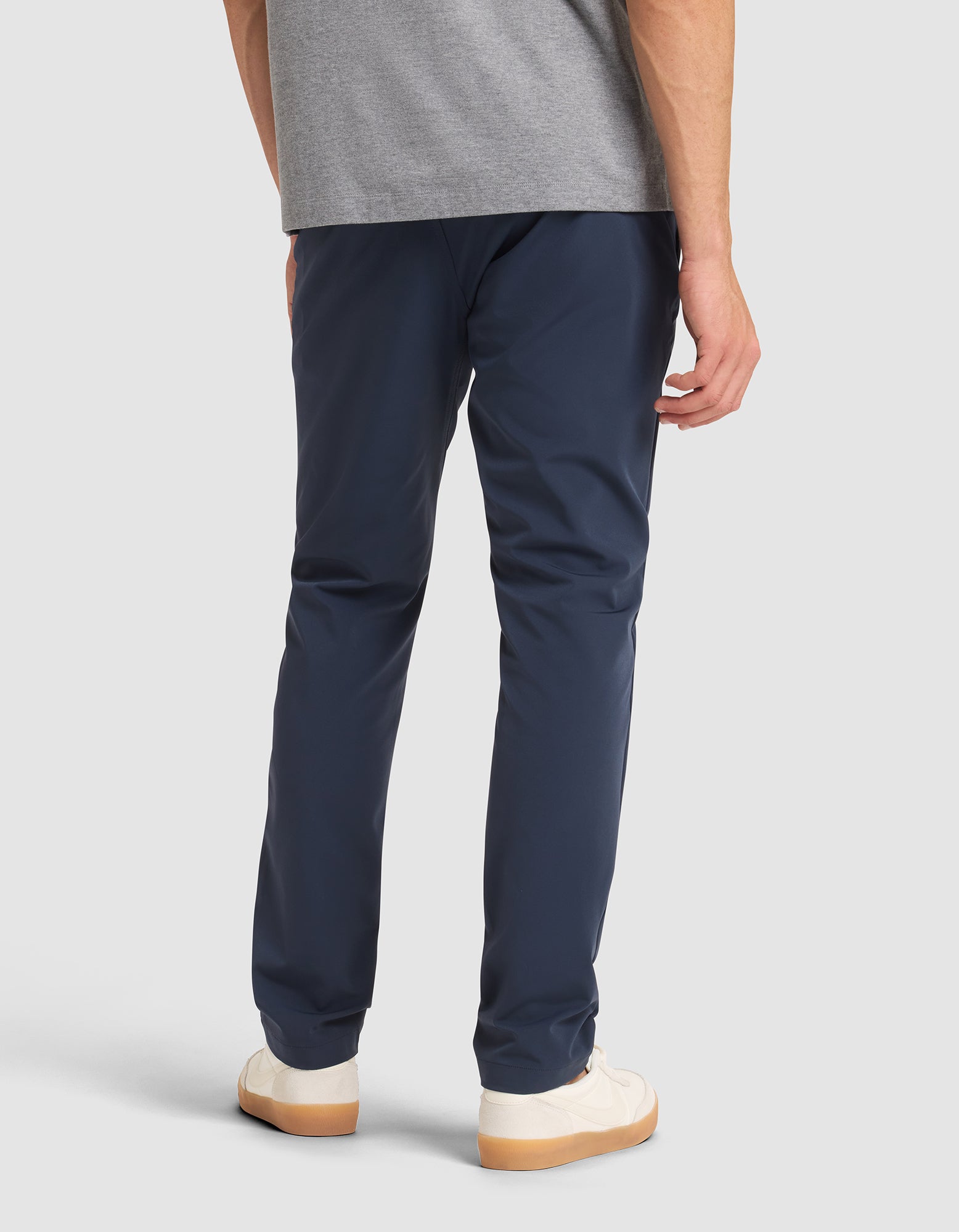 A person is shown from the back, standing against a plain background, wearing Cozy Earth’s Men's Everywhere Pant in navy blue, a gray t-shirt, and white sneakers with gum soles. 