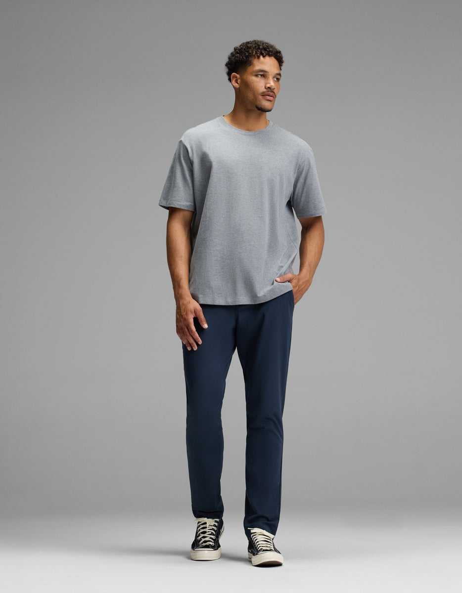 A man stands against a gray background, wearing the Cozy Earth Men’s Everywhere Pant in dark blue, paired with a light gray t-shirt and black-and-white sneakers. One hand is in his pocket as he looks slightly to the side with a neutral expression.