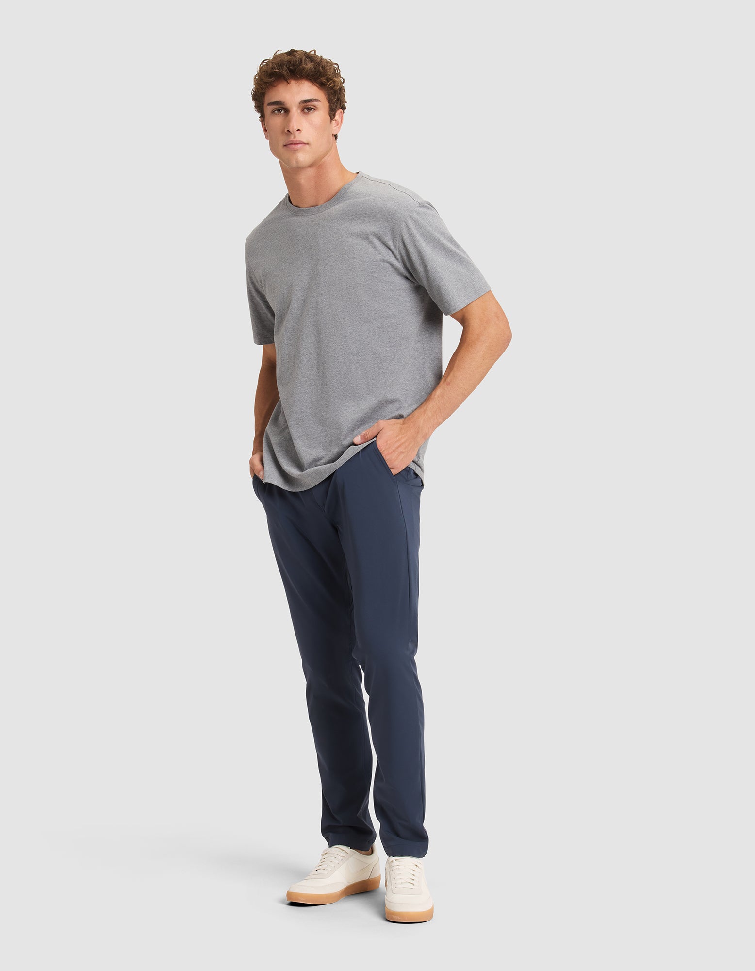 A man stands against a light gray background wearing Cozy Earth’s Men's Everywhere Pant in navy, a gray t-shirt, and white sneakers. He has short curly hair, hands in pockets, and looks slightly to the side. 