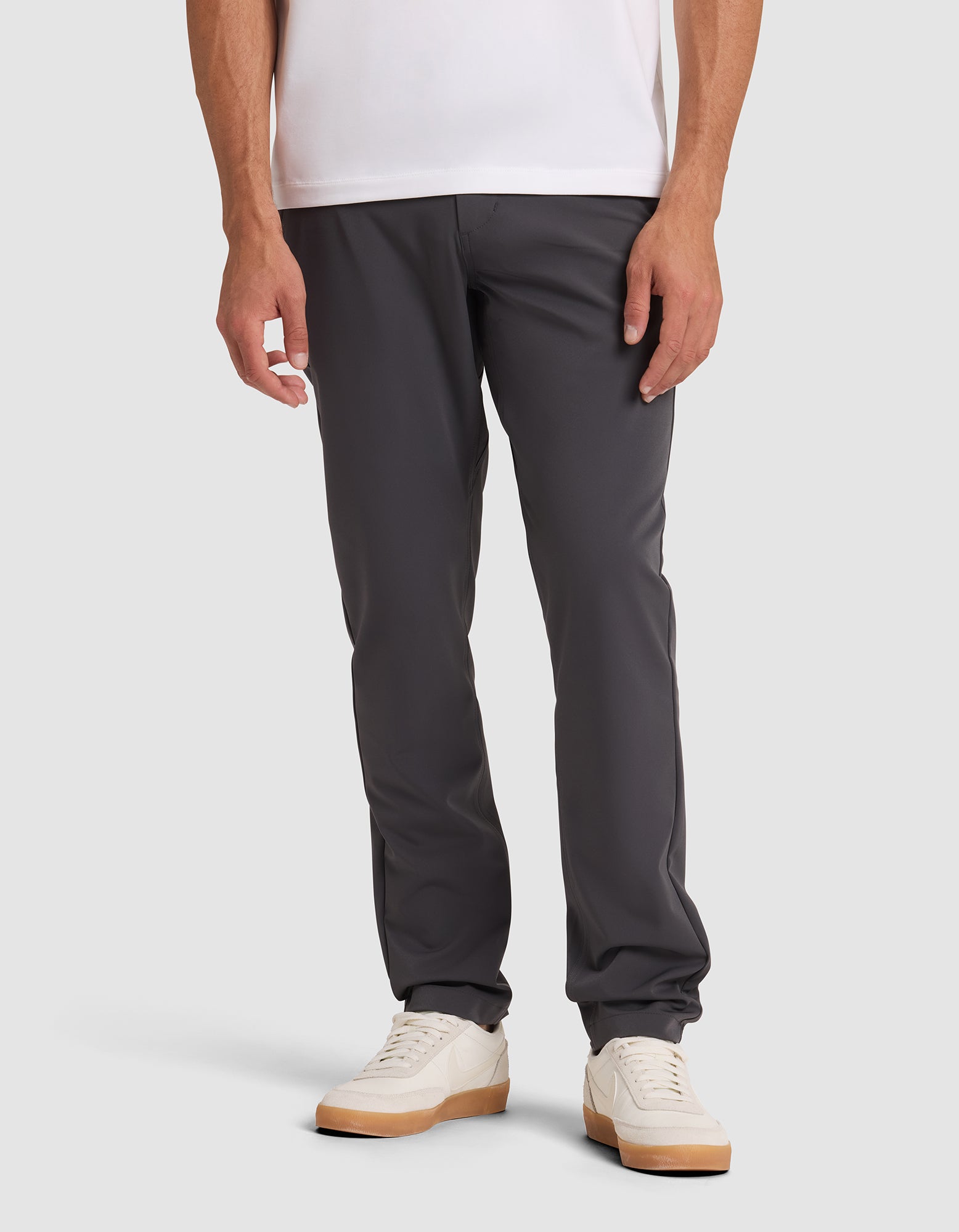 A person wearing Cozy Earth's Men's Everywhere Pant in dark gray stands against a plain light background, paired with a white t-shirt and white sneakers with tan soles. The image is cropped from the shoulders down.  