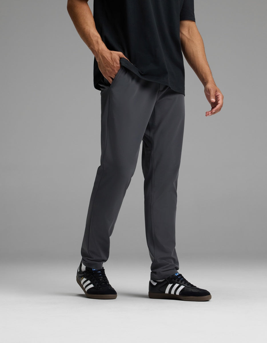 Wearing Cozy Earth’s Men’s Everywhere Pant in dark gray, along with a black T-shirt and black Adidas sneakers with white stripes, the individual stands against a plain gray background. Their right hand rests in their pocket, and their head is out of frame.