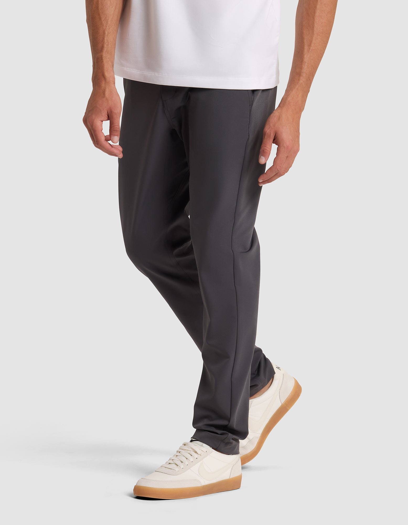 A person wearing Cozy Earth's Men's Everywhere Pant in dark gray, paired with a white shirt and white sneakers with tan soles, stands against a light gray background. Only the lower half of the body is shown. 