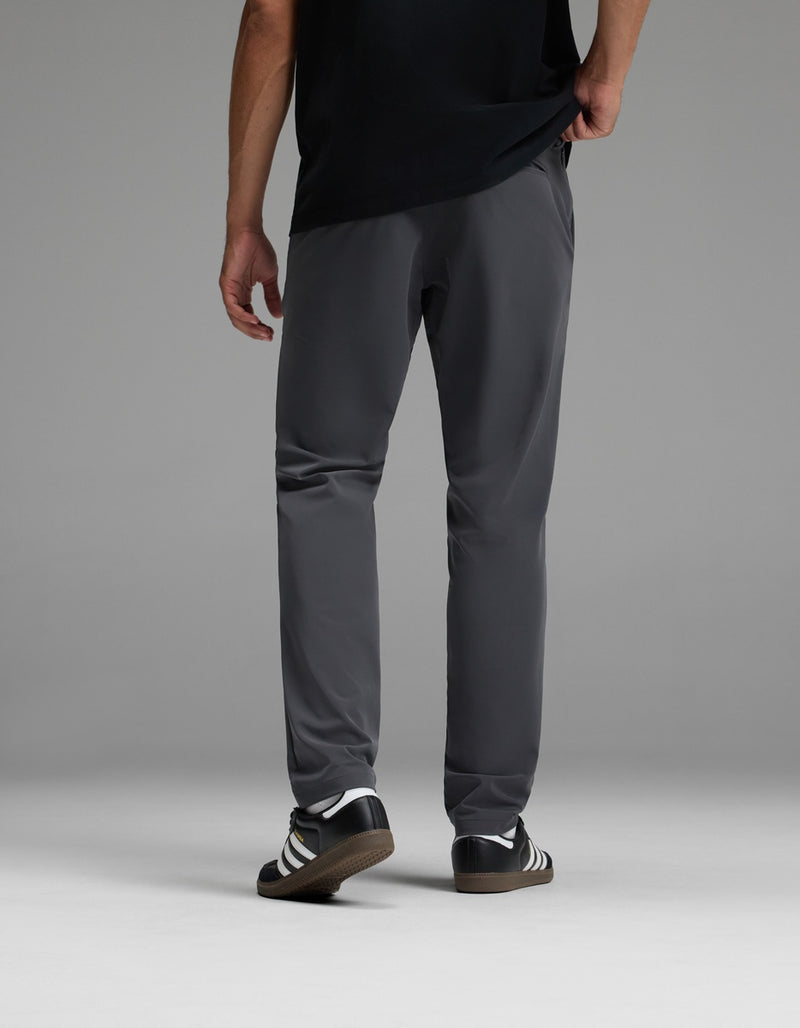 A person stands facing away, wearing a black shirt, Cozy Earth Men’s Everywhere Pant in gray, and black-and-white Adidas sneakers, against a plain gray background.