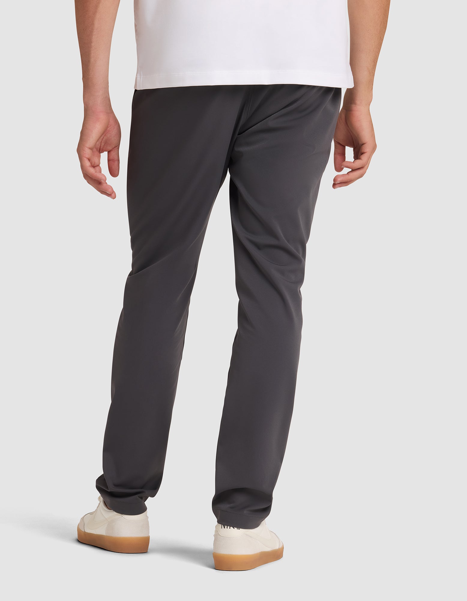 A person is seen from behind, standing against a light background, wearing Cozy Earth's Men's Everywhere Pant in dark gray, paired with a white shirt and white sneakers. 