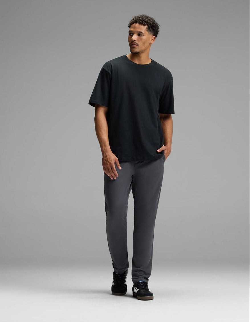A man stands against a plain gray background, wearing Cozy Earth’s Men’s Everywhere Pant in gray, paired with a black T-shirt and black shoes. He has short curly hair and is looking slightly to his right, with one hand in his pocket.