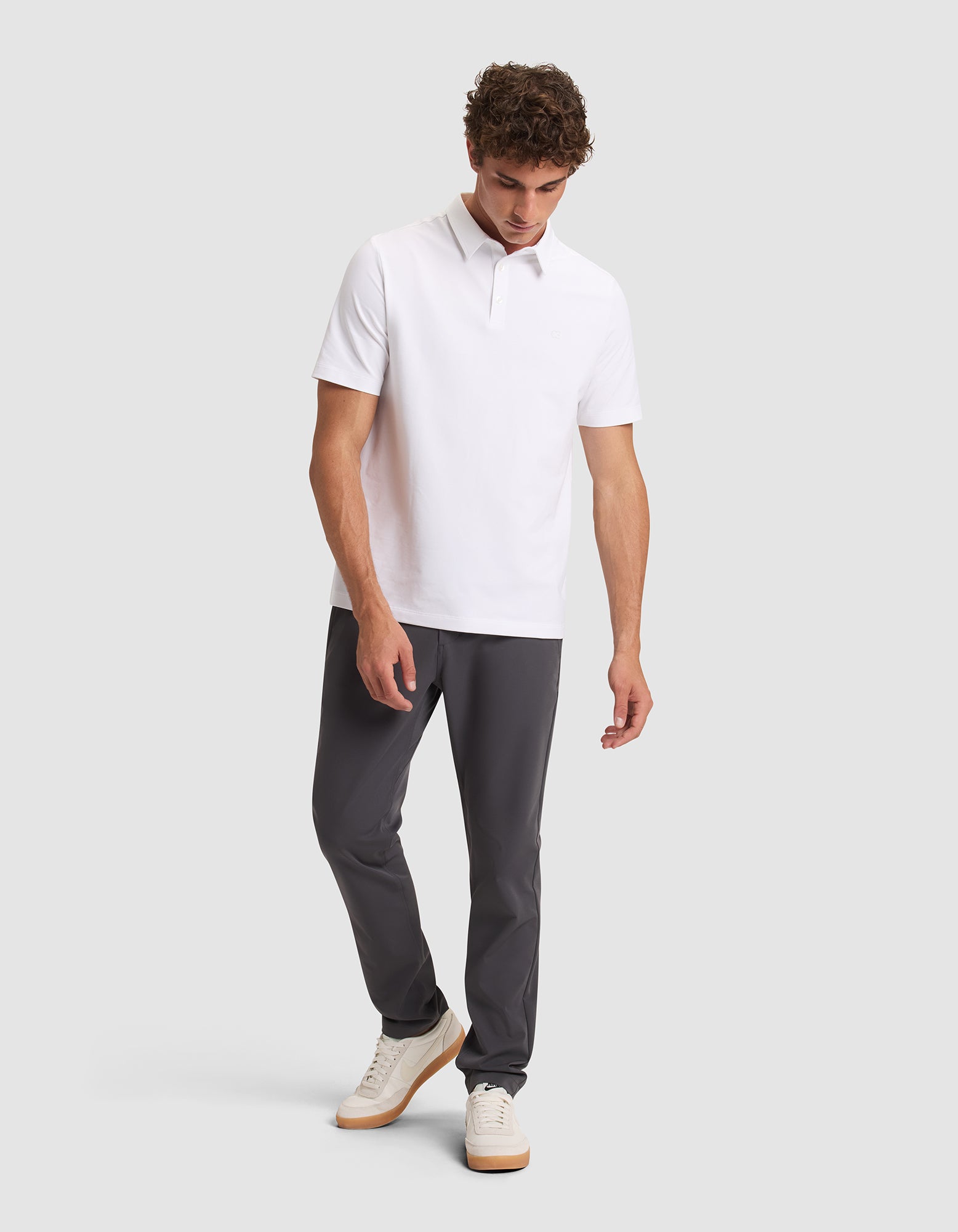 A man wearing Cozy Earth's Men's Everywhere Pant in dark gray, a white short-sleeve polo shirt, and white sneakers stands against a light gray background, looking down and slightly to the side. |Color:Graphite