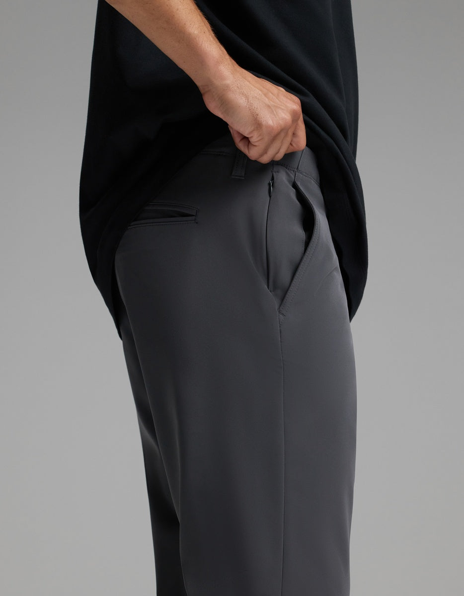 A person wearing Cozy Earth’s Men’s Everywhere Pant in dark gray and a black shirt lifts their shirt to reveal the side zipper pocket on the pants. The background is plain and gray.