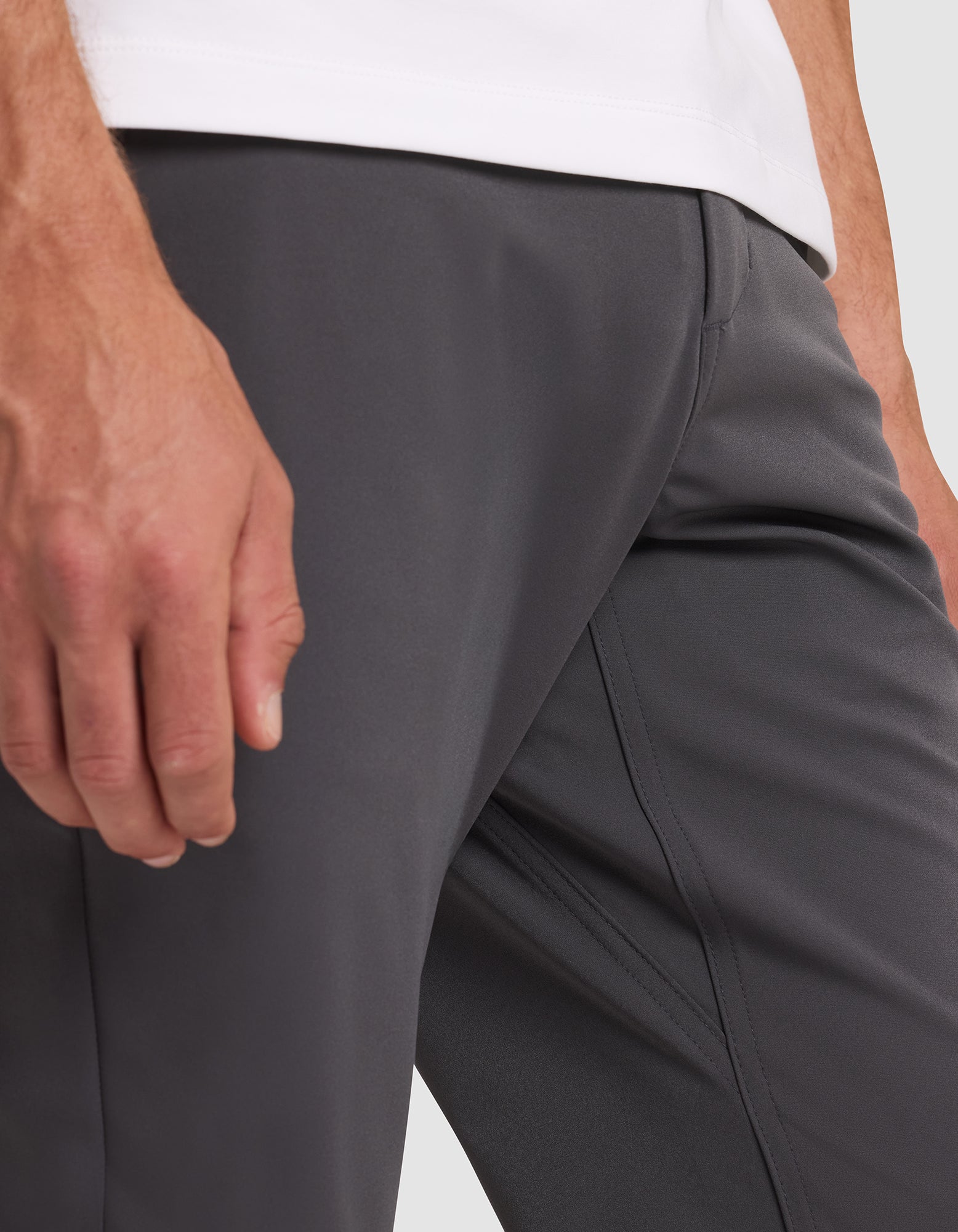 Close-up of a person wearing Cozy Earth's Men's Everywhere Pant in dark gray with a white shirt, their left hand resting by their thigh. Background is plain and minimal. 