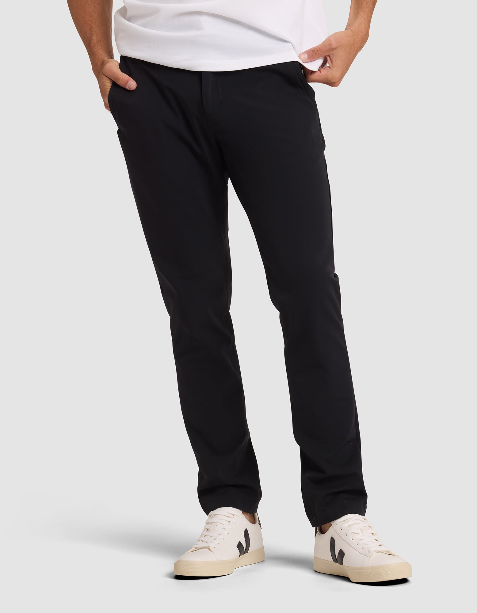 A person wearing Cozy Earth's Men's Everywhere Pant in black, a white t-shirt, and cream-colored sneakers with black detailing stands with hands in pockets against a plain light background. 