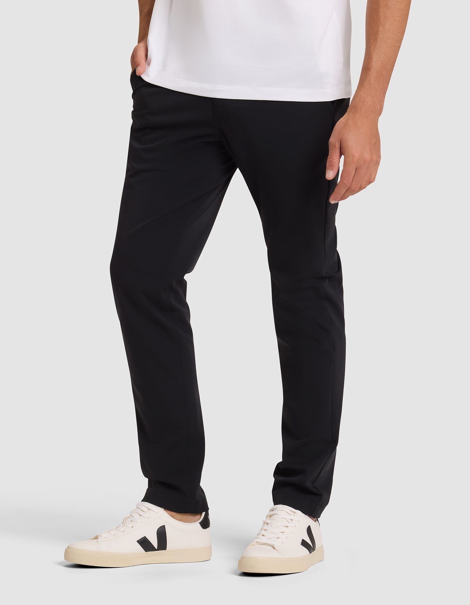 A person wearing Cozy Earth's Men's Everywhere Pant in black, a white t-shirt, and white sneakers with black detail stands against a plain light background, showing only the lower half of their body. 