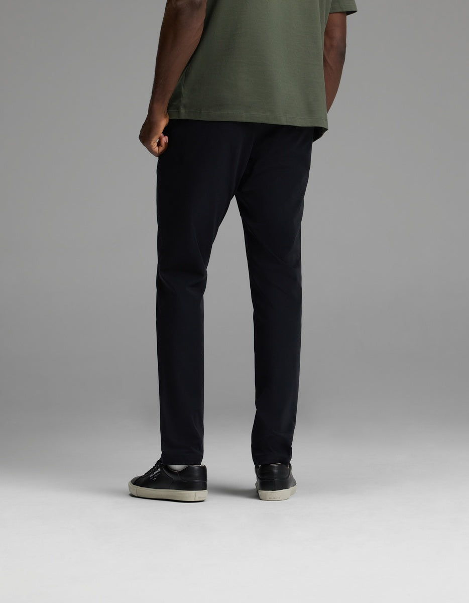 A person stands facing away against a plain gray background, wearing black Cozy Earth Men’s Everywhere Pants, paired with a green short-sleeve shirt and black sneakers with white soles.