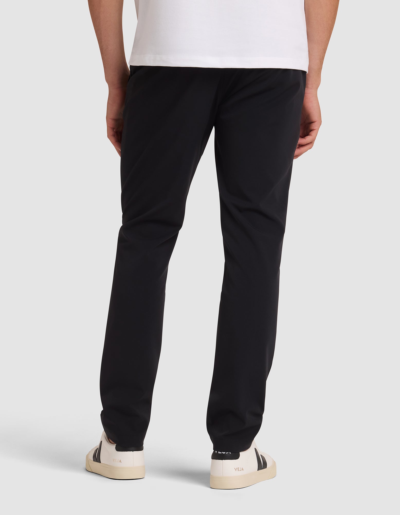 A person stands facing away against a plain light background, wearing Cozy Earth's Men's Everywhere Pant in black, a white t-shirt, and white sneakers with black detailing. 