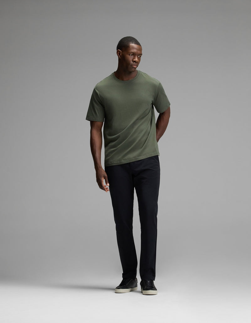 A man stands in a studio wearing Cozy Earth’s Men’s Everywhere Pant, paired with a green t-shirt and black shoes. He faces slightly to his left with one hand in his pocket against a plain gray background.