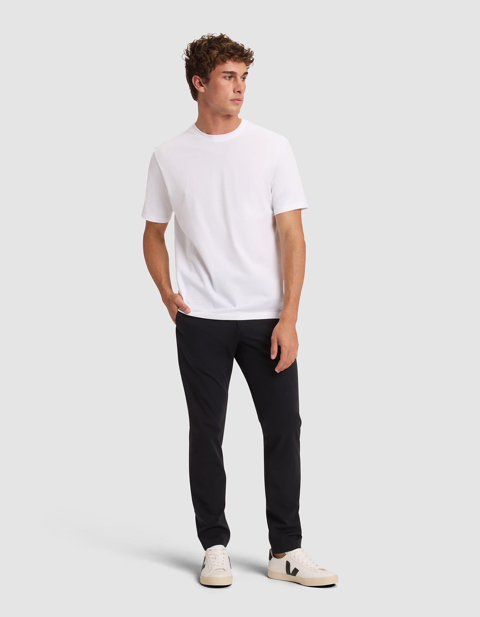 A man with short, curly hair stands against a plain background, looking left with one hand in his pocket. He wears Cozy Earth's Men's Everywhere Pant in black, a white t-shirt, and white sneakers with black details. 