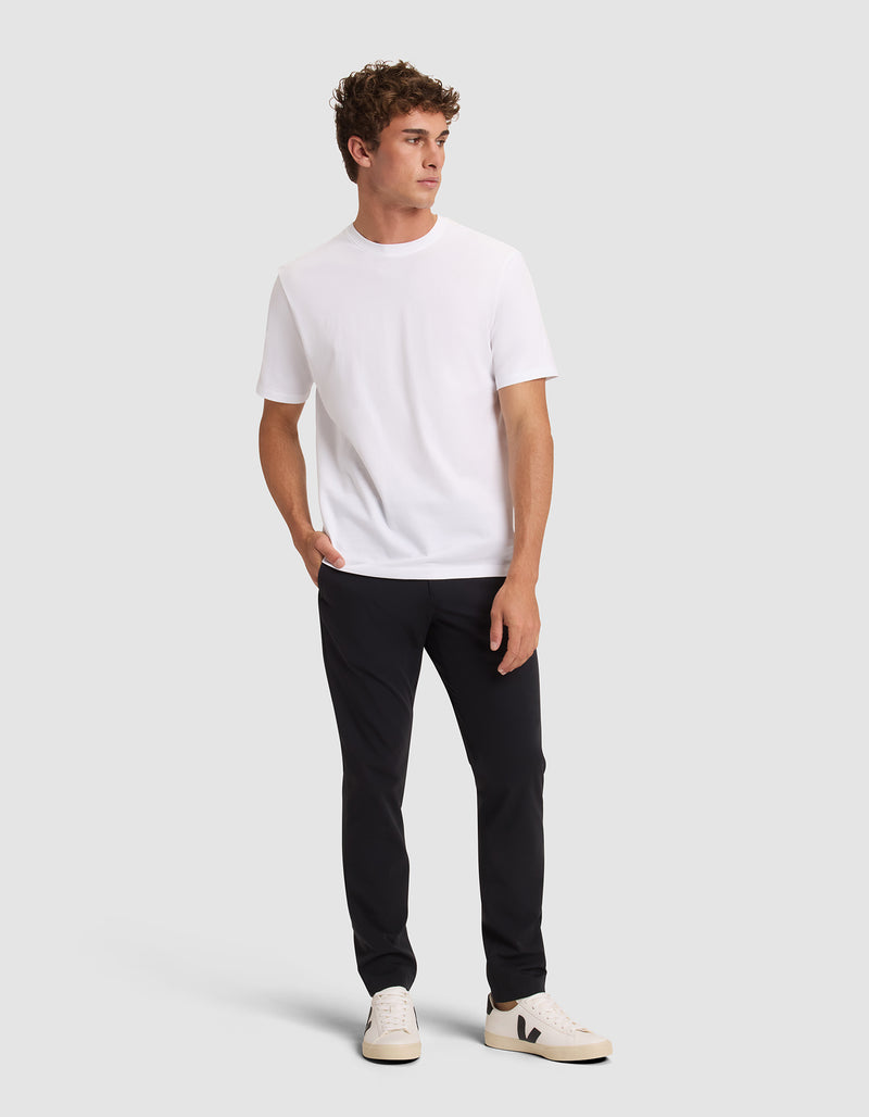 A man with short, curly hair stands against a plain background, looking left with one hand in his pocket. He wears Cozy Earth's Men's Everywhere Pant in black, a white t-shirt, and white sneakers with black details.