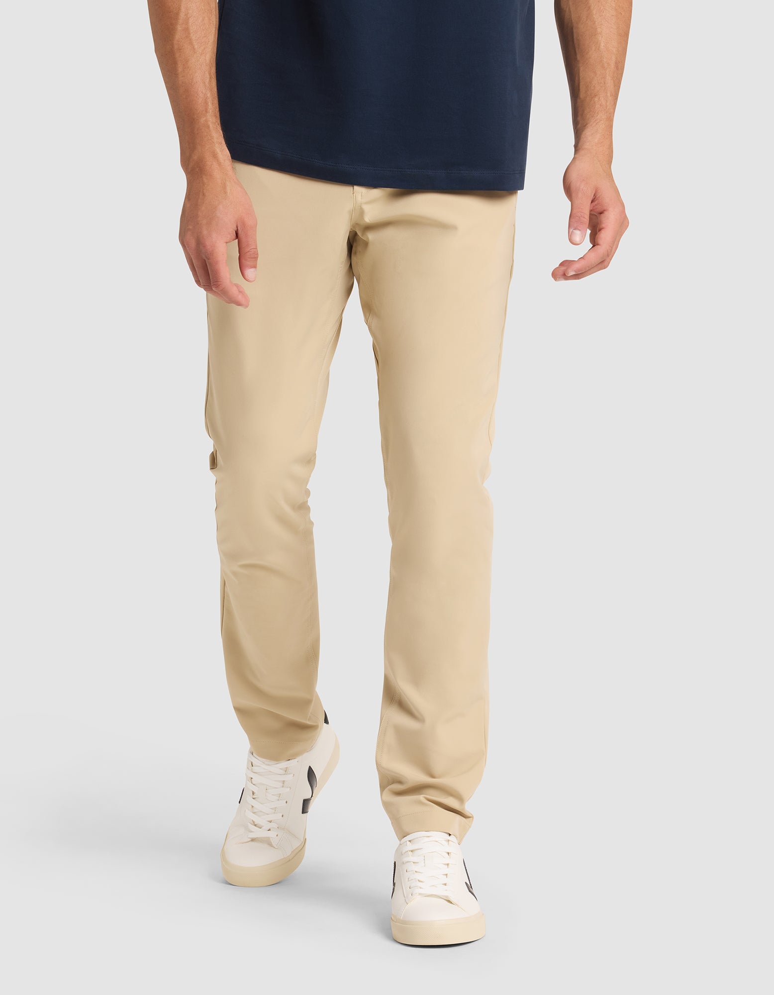 A model wears Cozy Earth's Men's Everywhere Pant in beige, paired with a navy shirt and white sneakers with black accents, standing against a plain light gray background. The image is cropped from the shoulders down. 