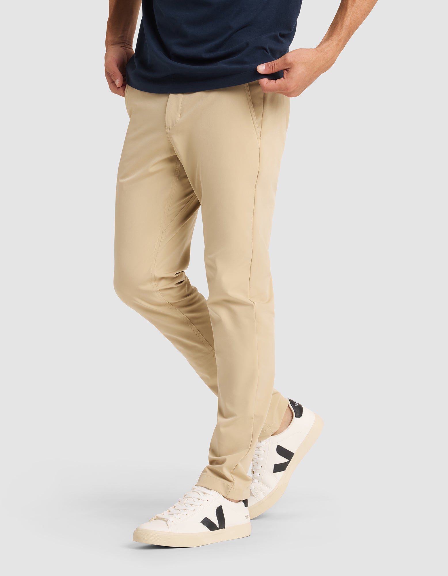 A person wearing Cozy Earth's Men's Everywhere Pant in beige, paired with a dark navy shirt and white sneakers with black "V" logos, stands hands-in-pockets against a plain light background. Only the lower half of the body is shown. 
