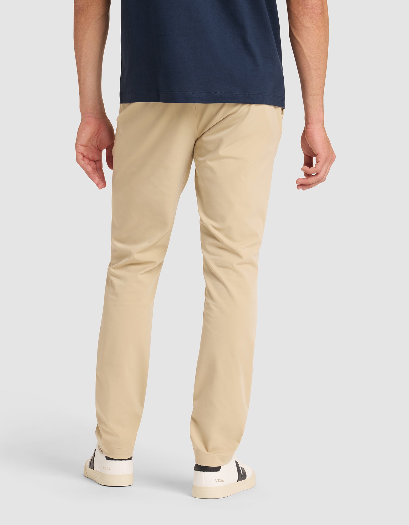 A person stands with their back to the camera, wearing Cozy Earth's Men's Everywhere Pant in beige, a navy shirt, and white sneakers with black accents against a plain light background. 