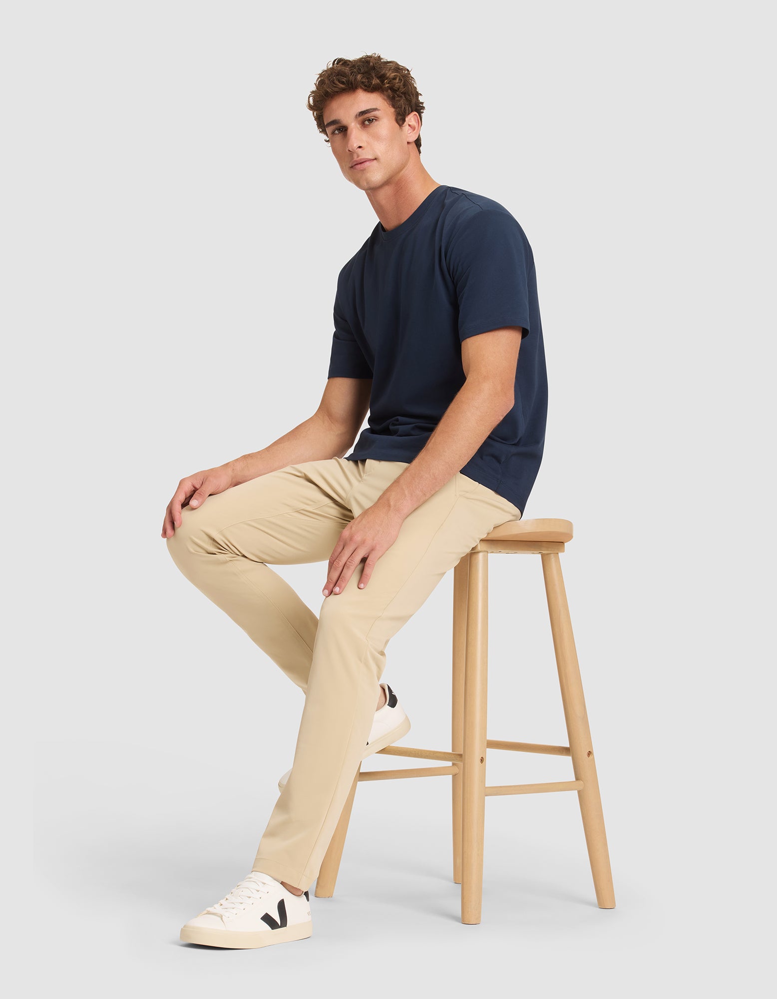 A young man sits on a wooden stool, wearing Cozy Earth's Men's Everywhere Pant in beige, a navy t-shirt, and white sneakers with a black "V" logo, against a plain light gray background. 