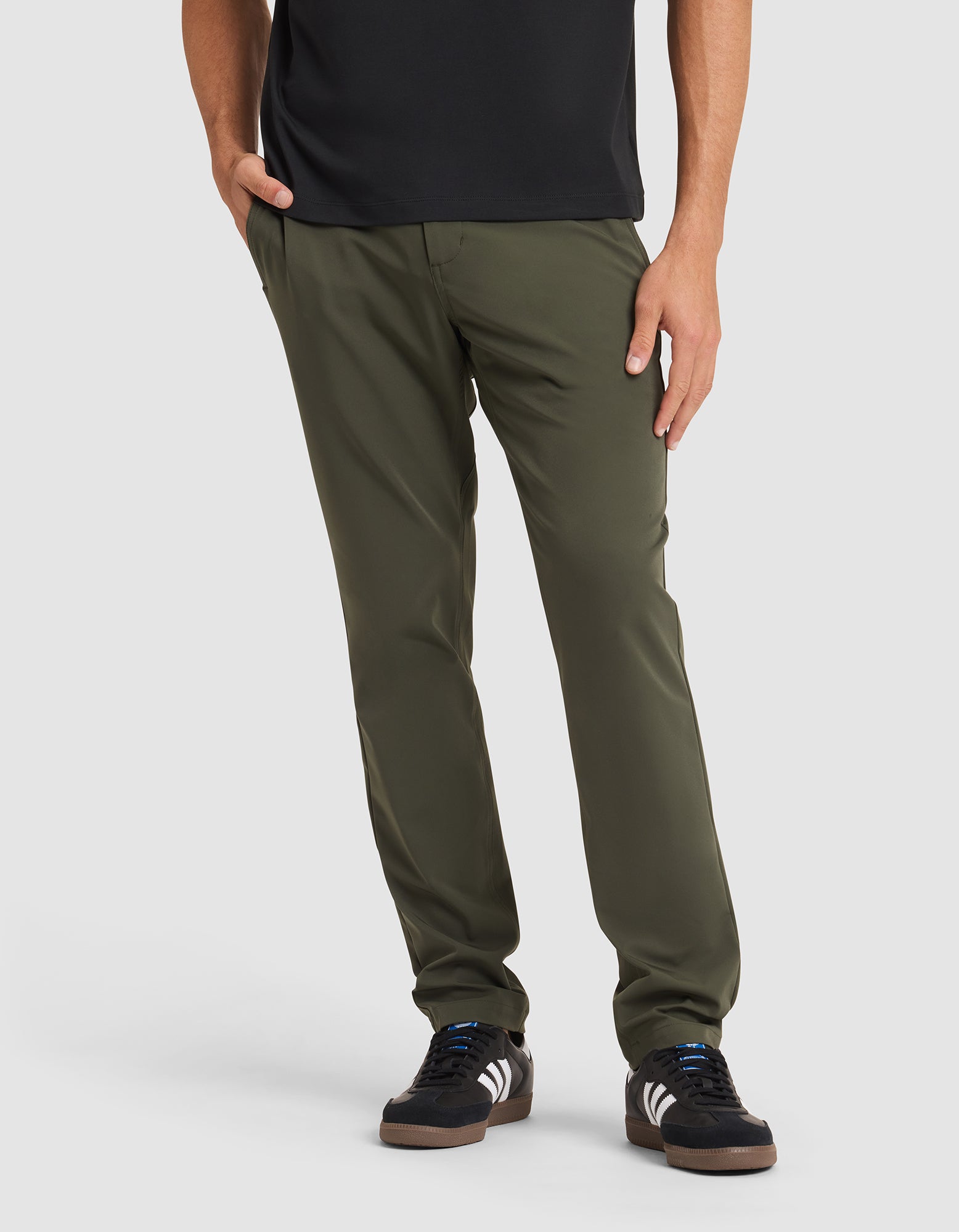 A person models Cozy Earth’s Men's Everywhere Pant in olive green, paired with a black shirt and black sneakers with white stripes, standing with one hand in their pocket against a plain white background. 