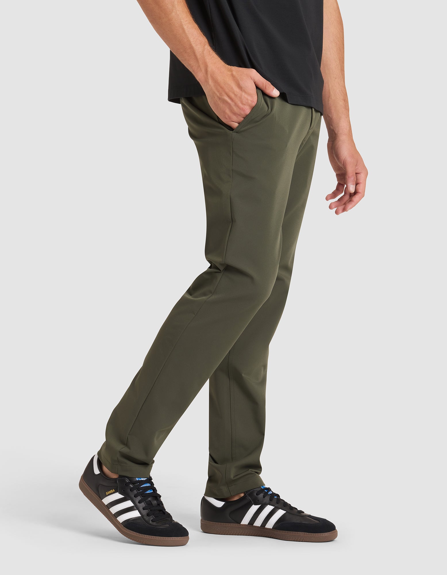 A person wearing Cozy Earth's Men's Everywhere Pant in olive green, a black shirt, and black Adidas sneakers with white stripes stands with one hand in their pocket against a plain white background. 