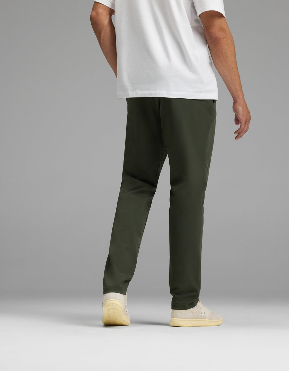 A man stands facing away against a gray background, wearing Cozy Earth’s Men’s Everywhere Pant in olive green, a white T-shirt, and off-white sneakers.