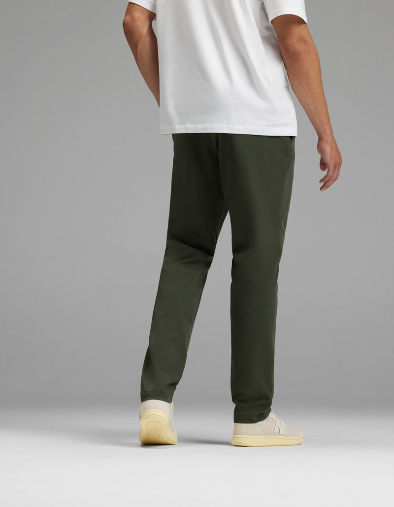 A man stands facing away against a gray background, wearing Cozy Earth’s Men’s Everywhere Pant in olive green, a white T-shirt, and off-white sneakers.