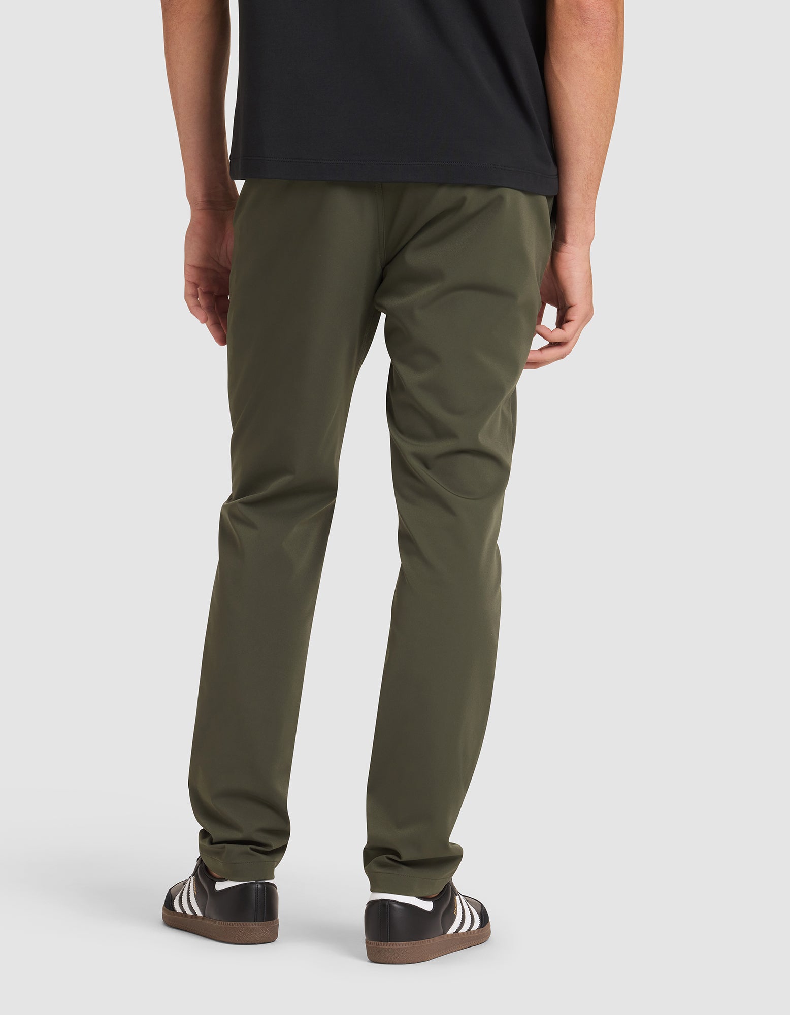 A person stands with their back to the camera, wearing Cozy Earth's Men's Everywhere Pant in olive green, a black shirt, and black sneakers with white stripes. 