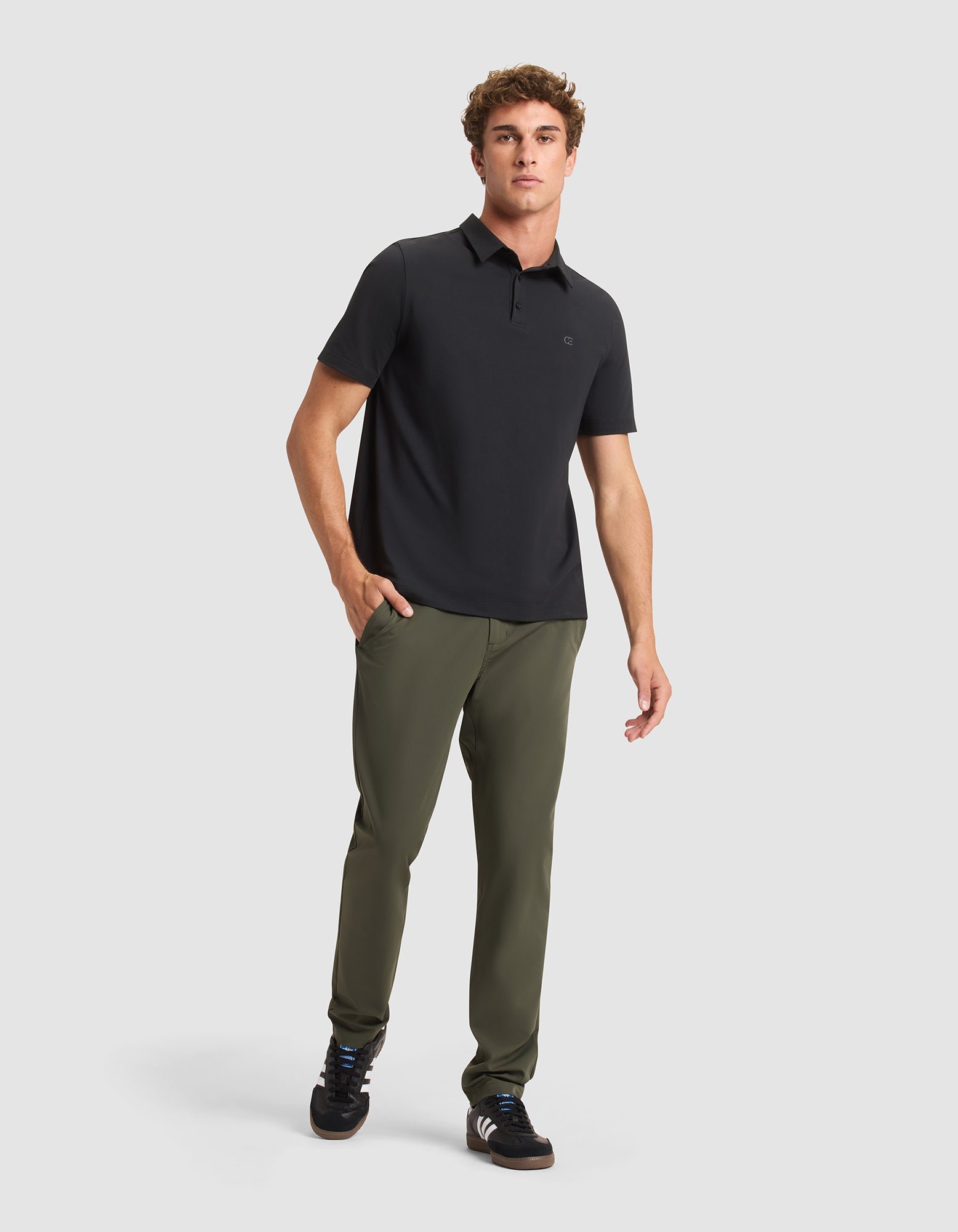 A man stands against a plain light background wearing Cozy Earth's Men's Everywhere Pant in olive green, paired with a black polo shirt and black sneakers. One hand is in his pocket and he has a neutral expression. 