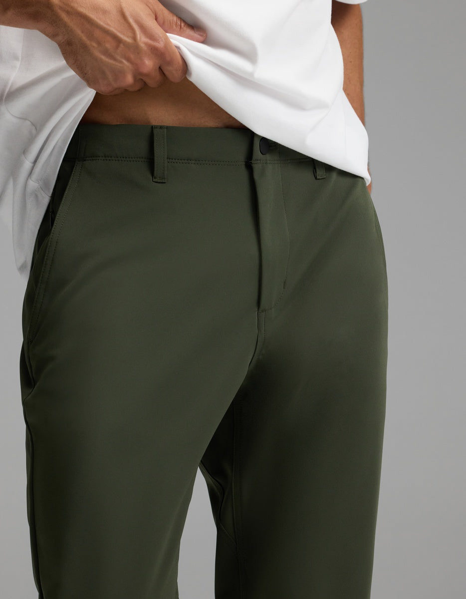 A person models the Cozy Earth Men’s Everywhere Pant in olive green, paired with a white shirt. They stand against a plain gray background, lifting the shirt slightly to reveal part of their midriff.