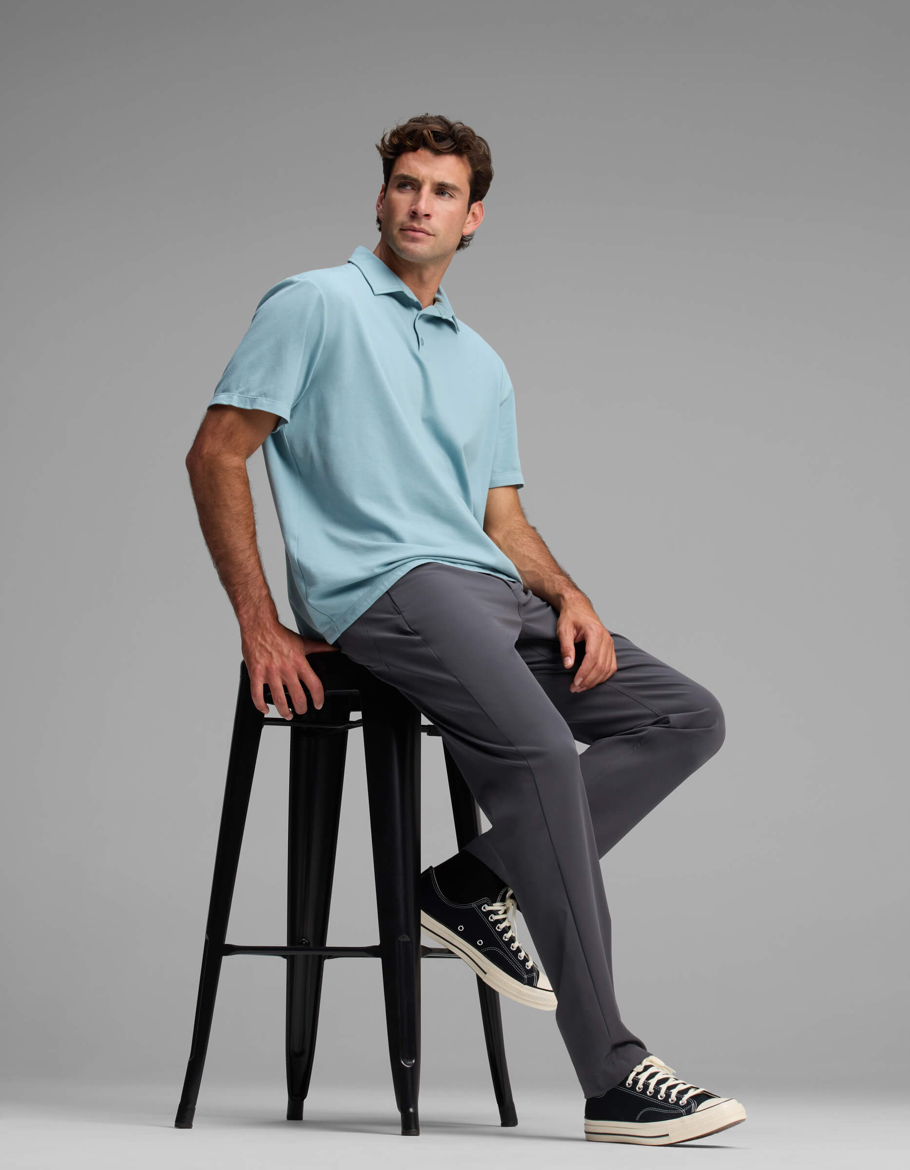 A man in Cozy Earth's Men's Everywhere Pant - Relaxed Fit, paired with a light blue polo and black sneakers, sits on a tall black stool against a plain gray background, gazing off to the side.