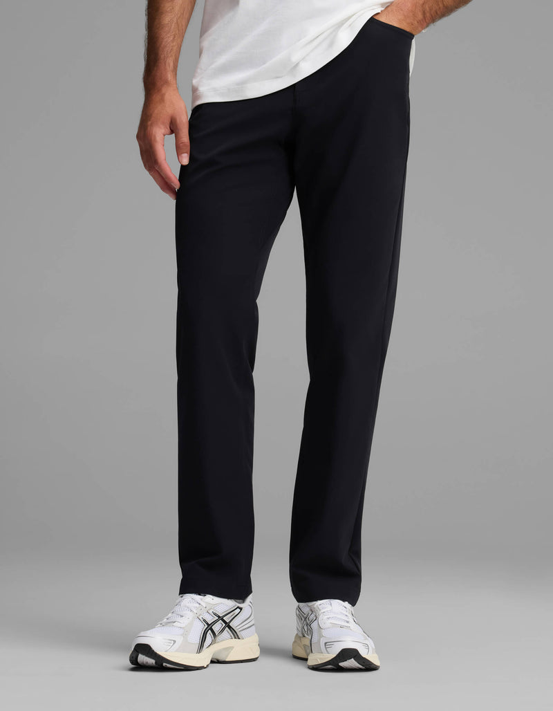 A person wearing Cozy Earth's Men's Everywhere Pant - Relaxed Fit in black, a white T-shirt, and white sneakers stands with one hand in their pocket against a plain gray background.