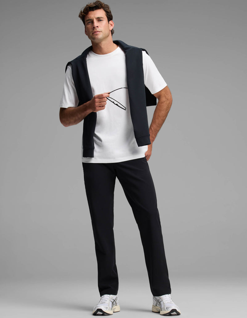 A man poses against a gray background wearing Cozy Earth's Men's Everywhere Pant - Relaxed Fit, a white T-shirt, white sneakers, and a dark jacket over his shoulders, holding glasses in one hand.