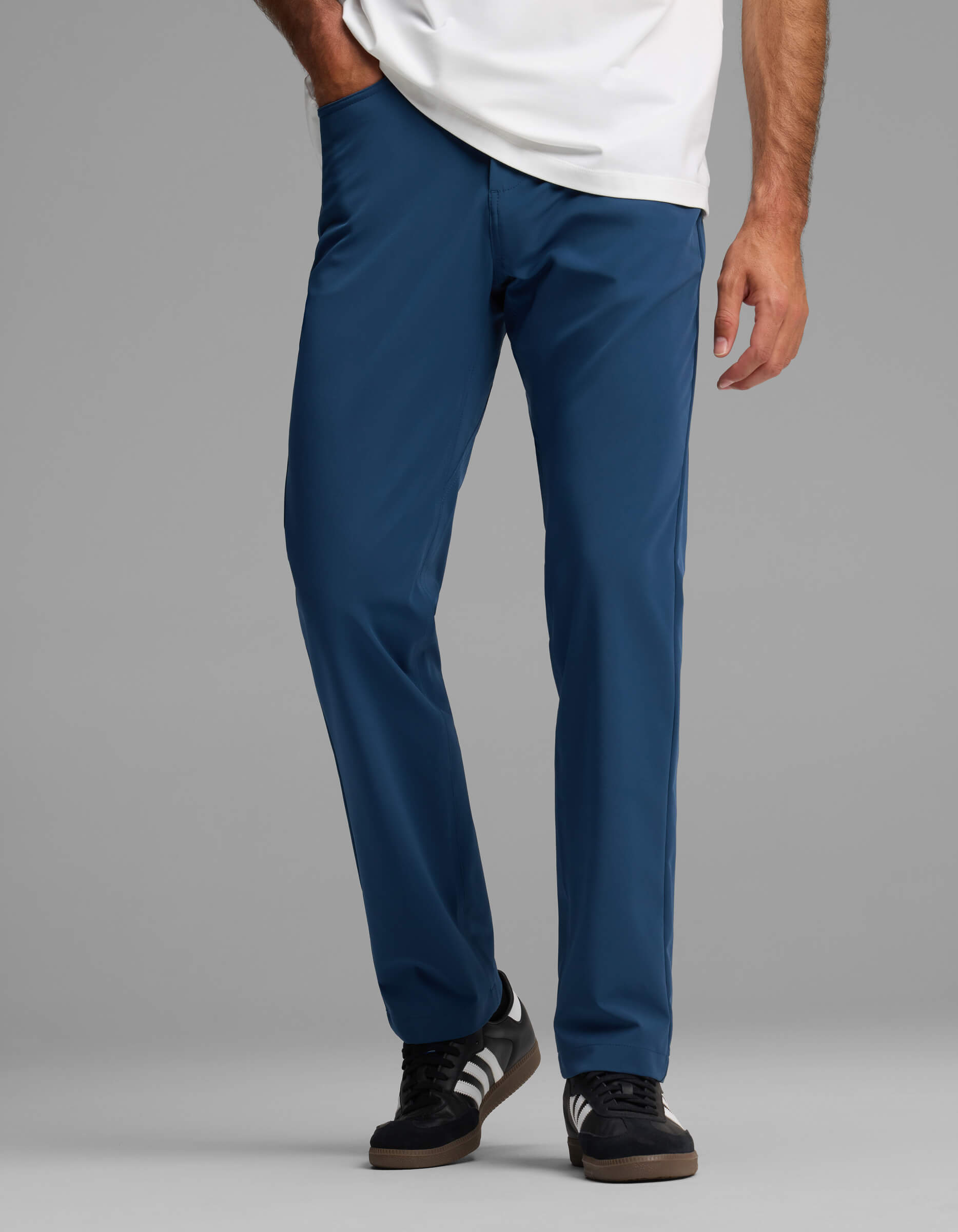 A person wearing Cozy Earth Men's Everywhere Pant - Relaxed Fit in blue, paired with a white shirt and black sneakers with white stripes, stands against a plain gray background.