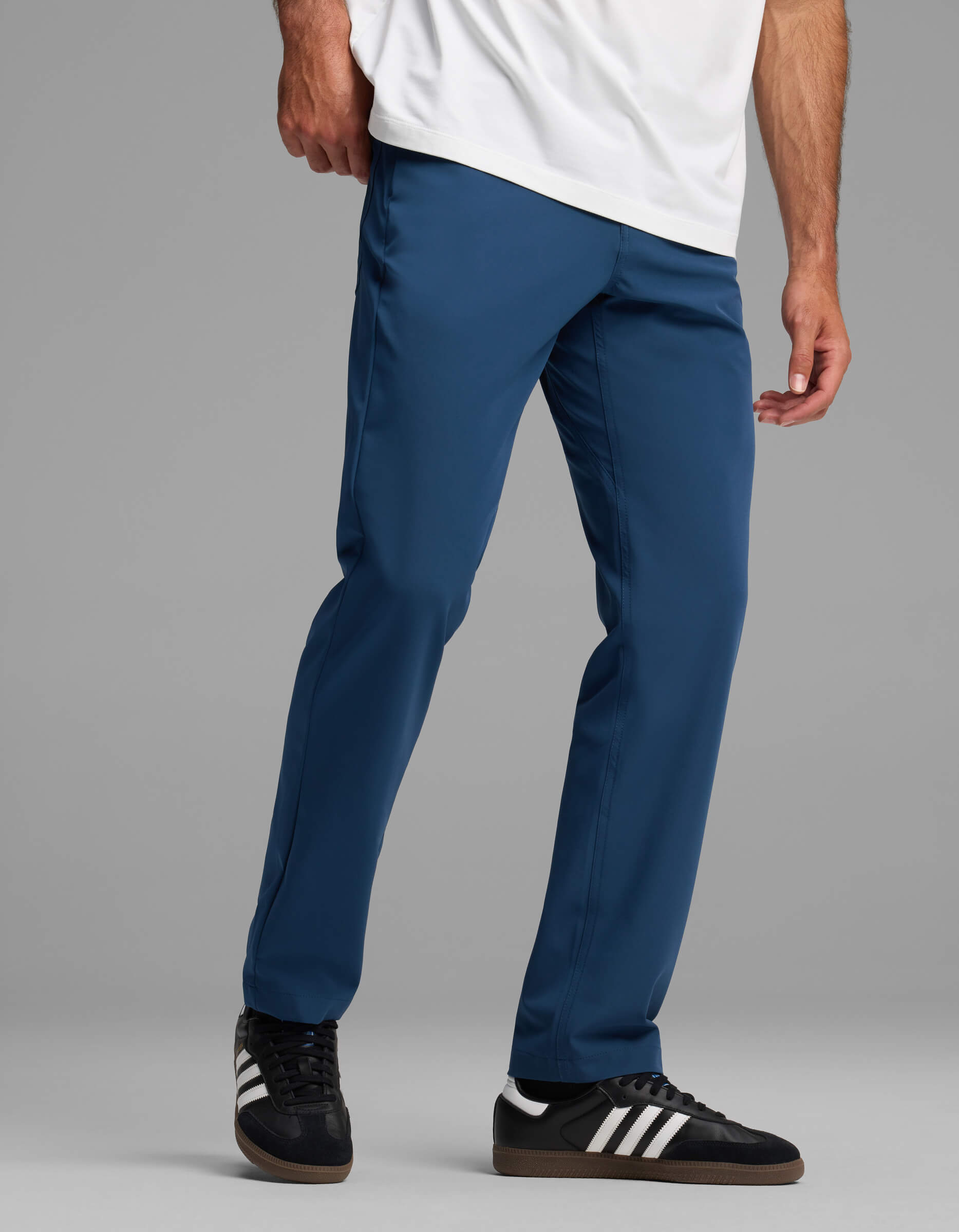 A person models Cozy Earth's Men's Everywhere Pant - Relaxed Fit in blue with a white shirt and black Adidas sneakers, standing against a gray background. The image is cropped at the shoulders.