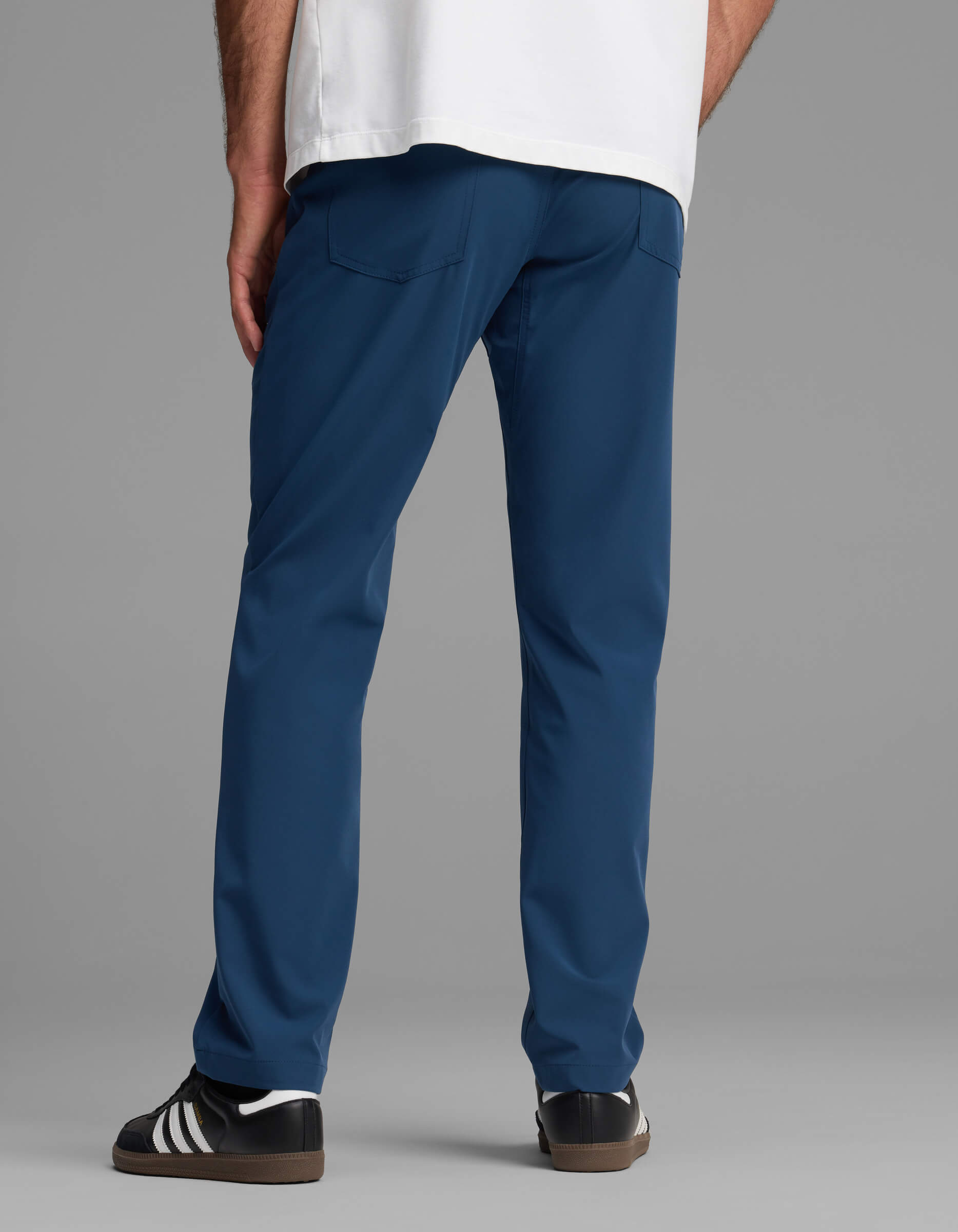 A person stands facing away against a plain gray background, wearing Cozy Earth's Men's Everywhere Pant - Relaxed Fit in blue, paired with a white shirt and black sneakers with white stripes.