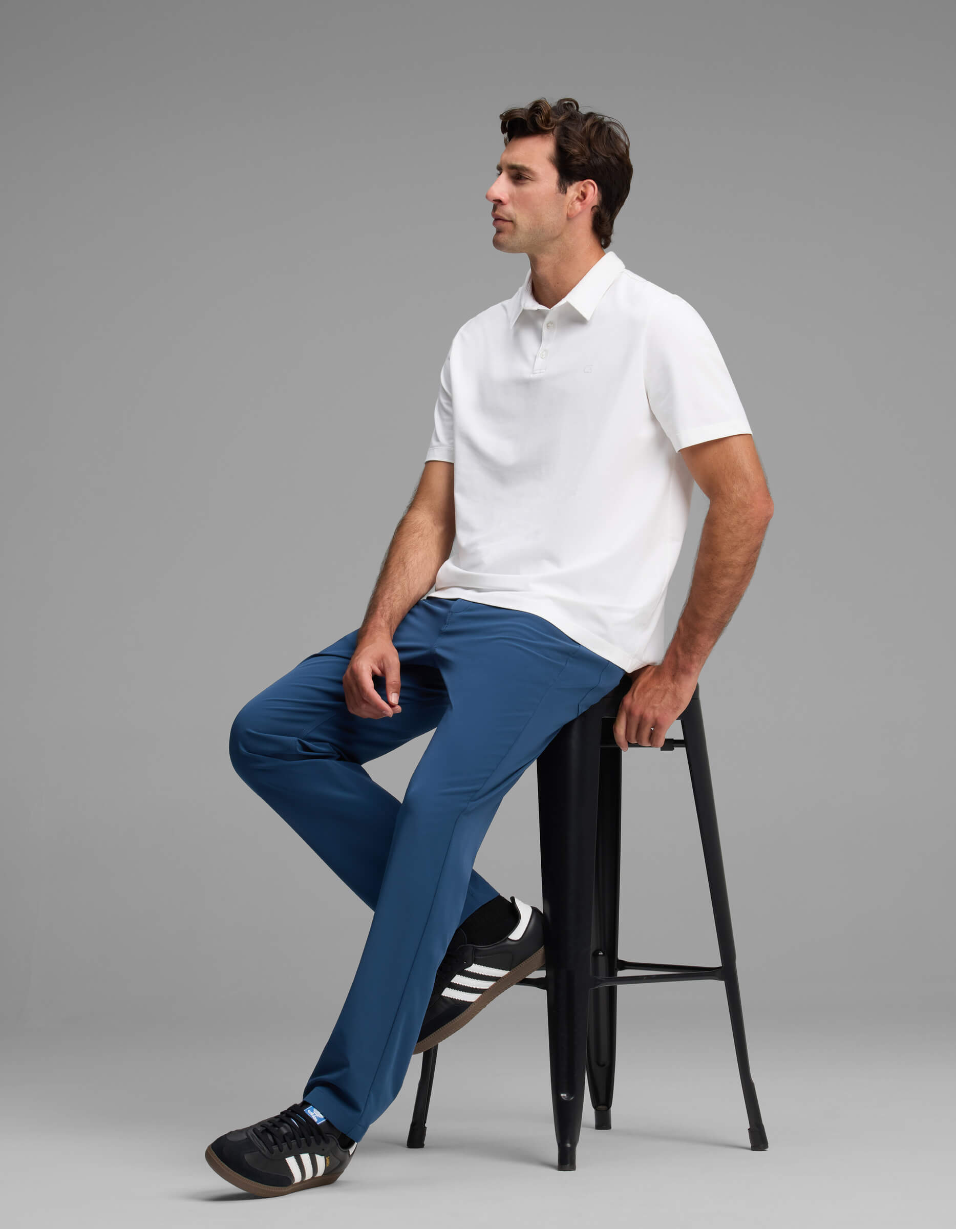 A man sits on a tall black stool against a plain gray background, wearing Cozy Earth's Men's Everywhere Pant - Relaxed Fit with a white polo shirt and black Adidas sneakers, looking to the side with a relaxed expression.