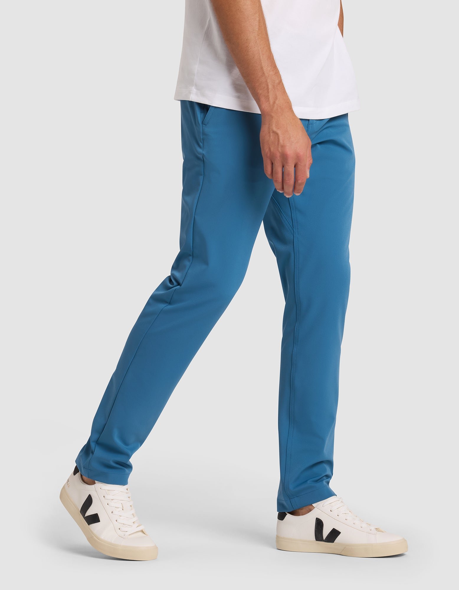 A person wearing Cozy Earth's Men's Everywhere Pant in teal-blue, a white t-shirt, and white sneakers with black "V" logos walks against a plain light gray background.  