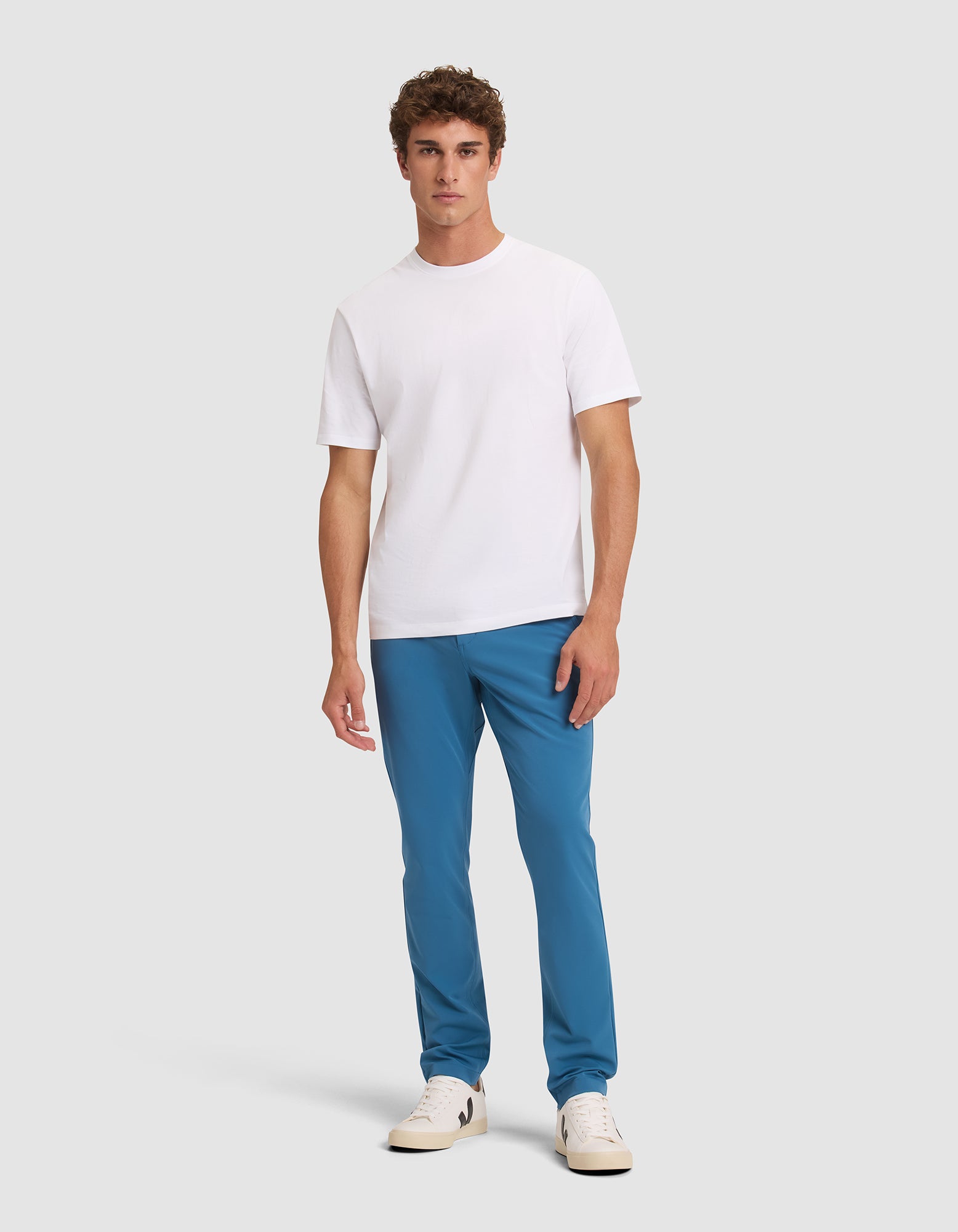A man stands against a light gray background, wearing Cozy Earth's Men's Everywhere Pant in blue, a plain white short-sleeve t-shirt, and white sneakers. He faces forward with a neutral expression and relaxed posture.  