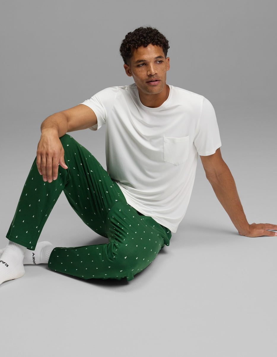 A man sits on the floor wearing a white pocket t-shirt and Cozy Earth Men’s First Layer Pajama Pant in green with a small white tree pattern.