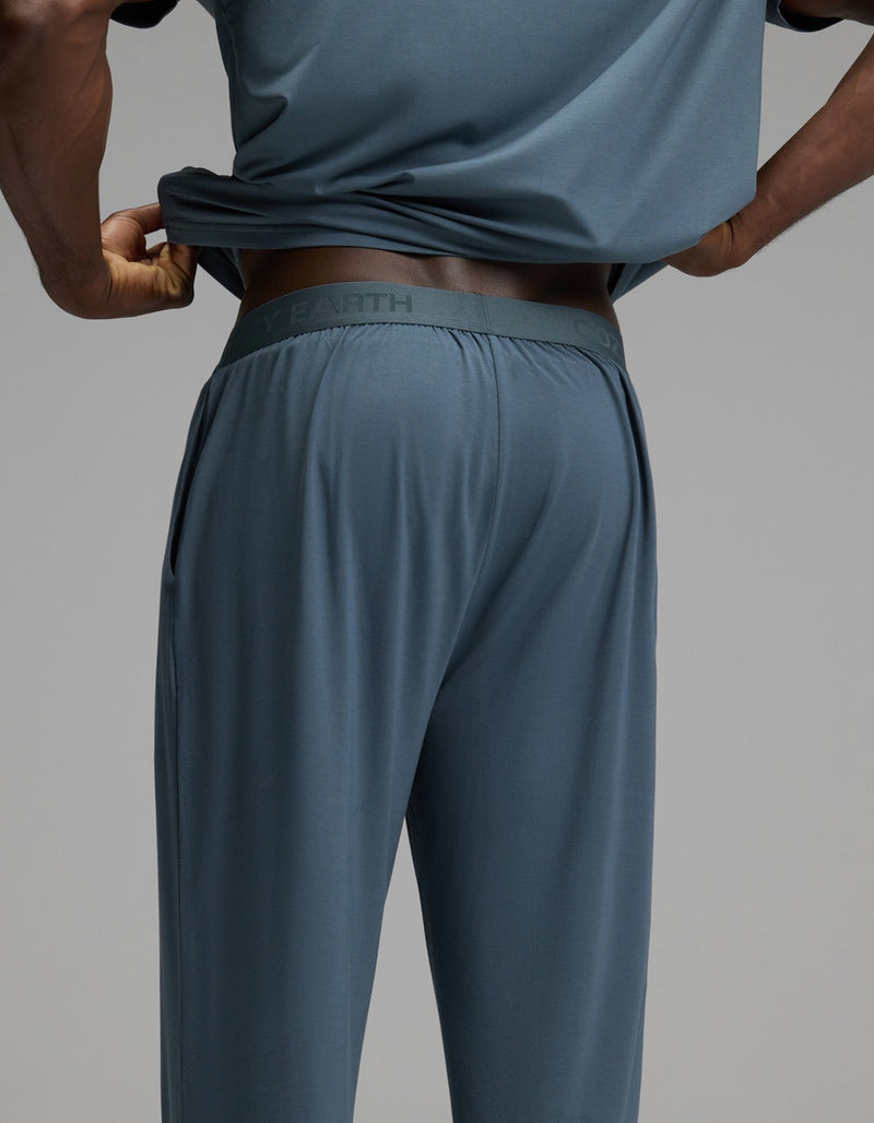 A person is wearing the Cozy Earth Men’s First Layer Pajama Pant, paired with a blue-gray shirt. They are lifting their shirt slightly to reveal the waistband of the pajama pants. The background is plain gray.