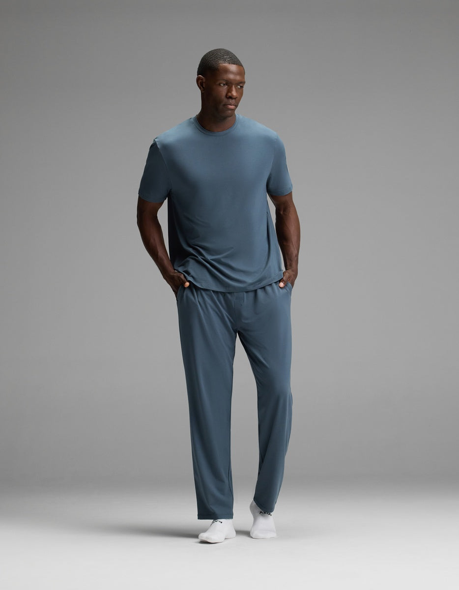 A man stands with his hands in his pockets, wearing the Cozy Earth Men’s First Layer Pajama Pant in blue paired with a matching t-shirt and white shoes, set against a plain gray background.|Color:Dawn