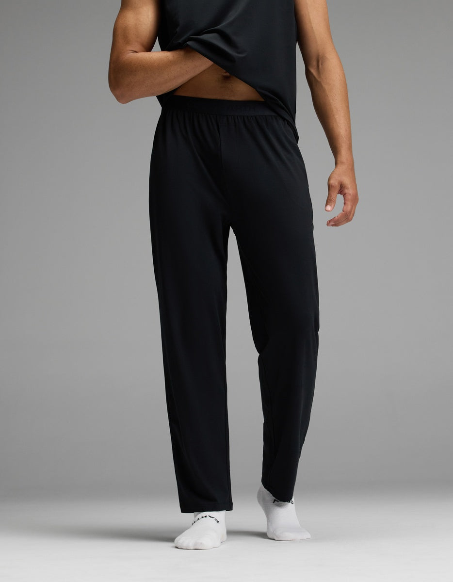 A person is shown from the lower body and part of one arm, standing against a gray background. They are wearing Cozy Earth’s Men’s First Layer Pajama Pant in black, paired with a black shirt and white socks. The person lifts their shirt slightly to reveal their midsection.|Color:Jet Black