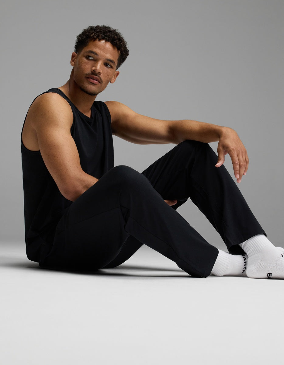A man wearing Cozy Earth’s Men’s First Layer Pajama Pant sits on the floor with one leg bent and one arm resting on his knee, looking to the side against a plain gray background.