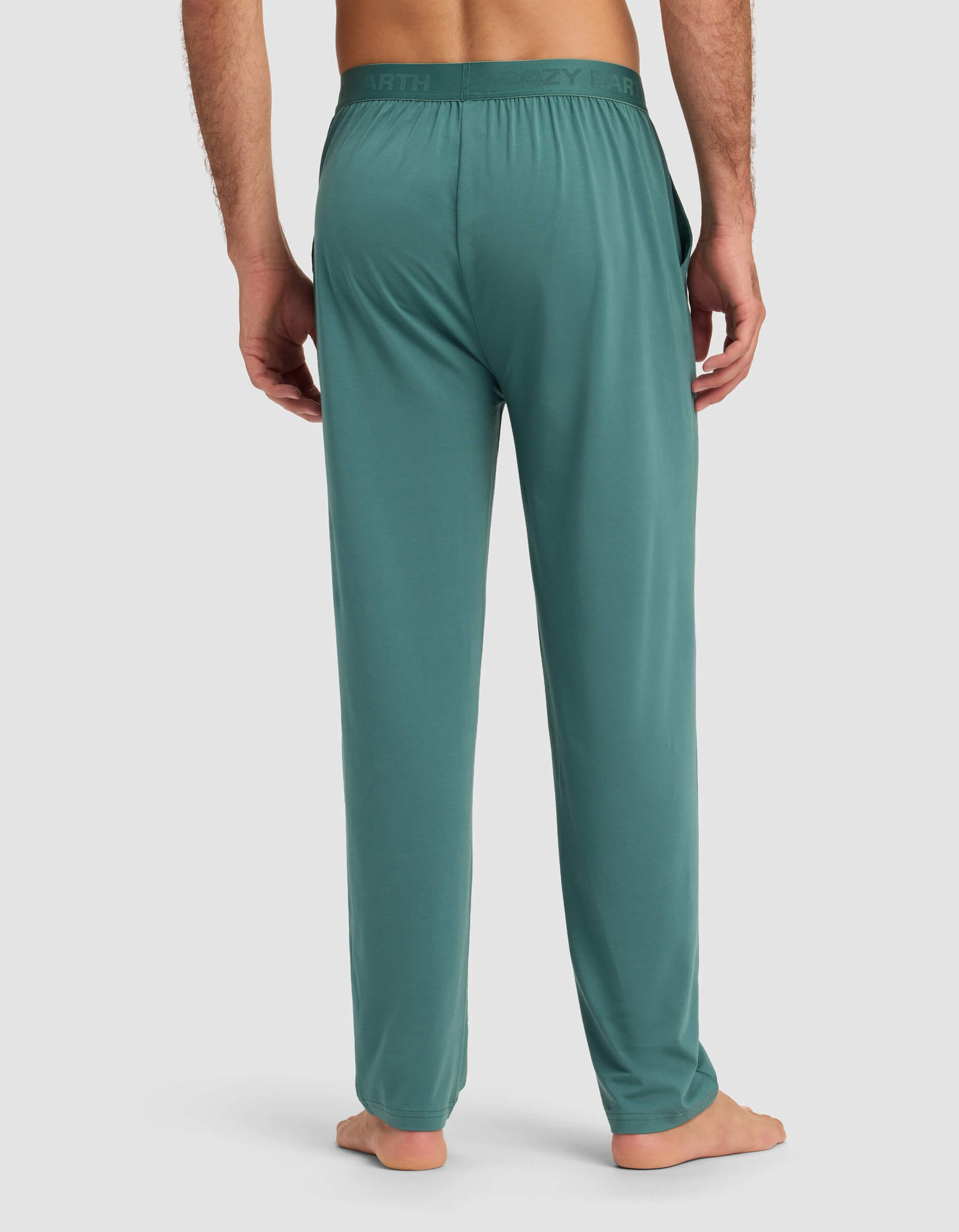 A man stands barefoot, back facing the camera, in Cozy Earth Men's First Layer Pajama Pant. The relaxed-fit teal pants feature side pockets and a subtly branded waistband, set against a plain light gray background.